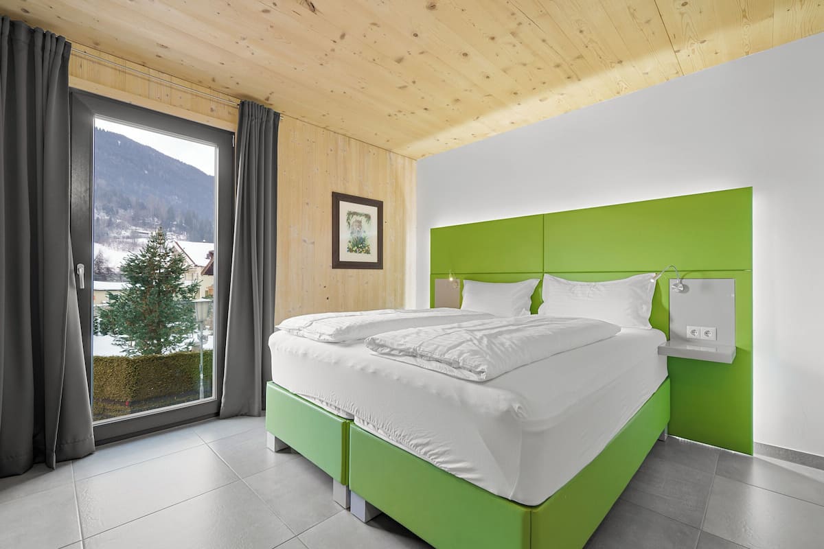 A modern bedroom with a green headboard, white bedding, and a view of a snowy landscape through a large window.