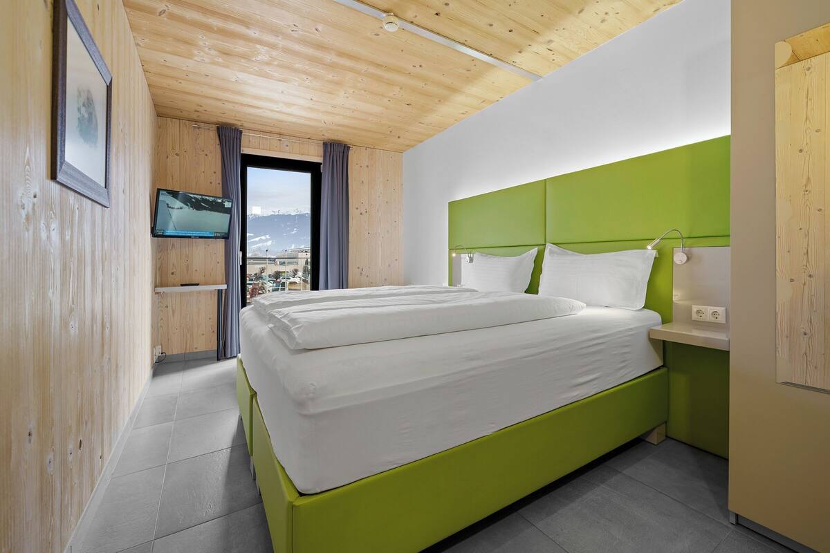 A hotel room with a bed, a TV, a window with curtains, and a view of mountains.
