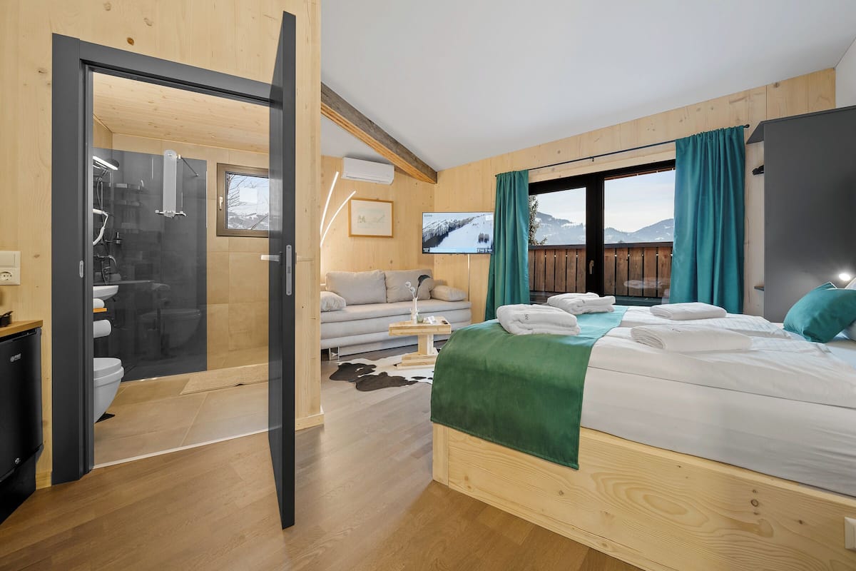 A modern hotel room with a bed, a sofa, a TV, and a view of mountains.