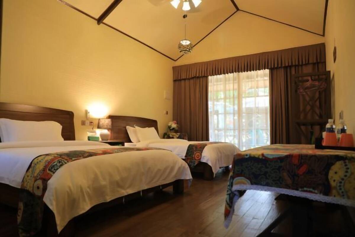 A hotel room with two beds, wooden flooring, a large window with curtains, and a ceiling fan.