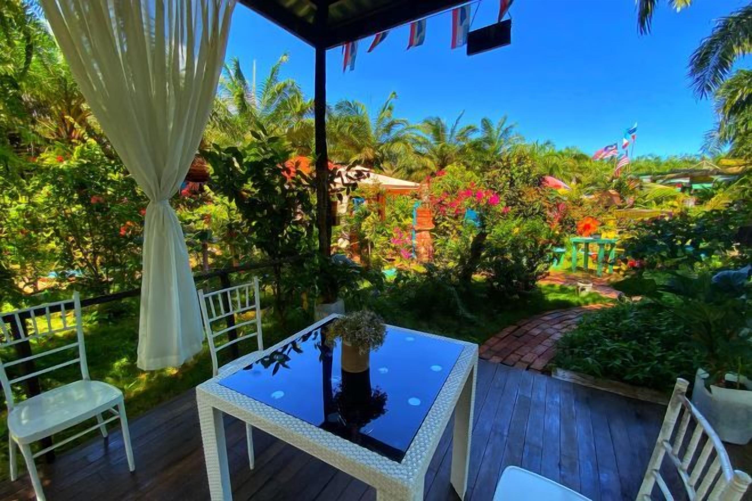 An outdoor patio with a table and chairs, surrounded by lush greenery and a clear blue sky.
