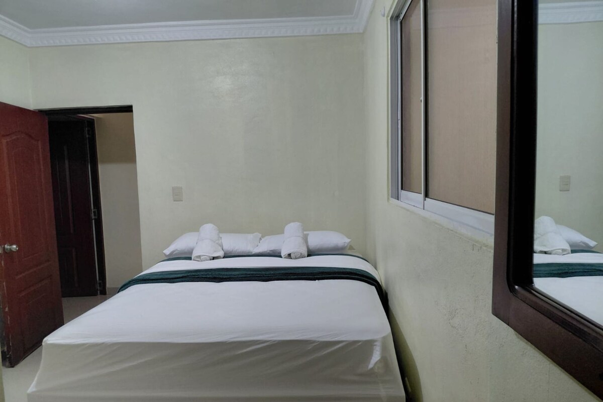 A hotel room with a bed, a wooden nightstand, a window, and a door leading to another room.