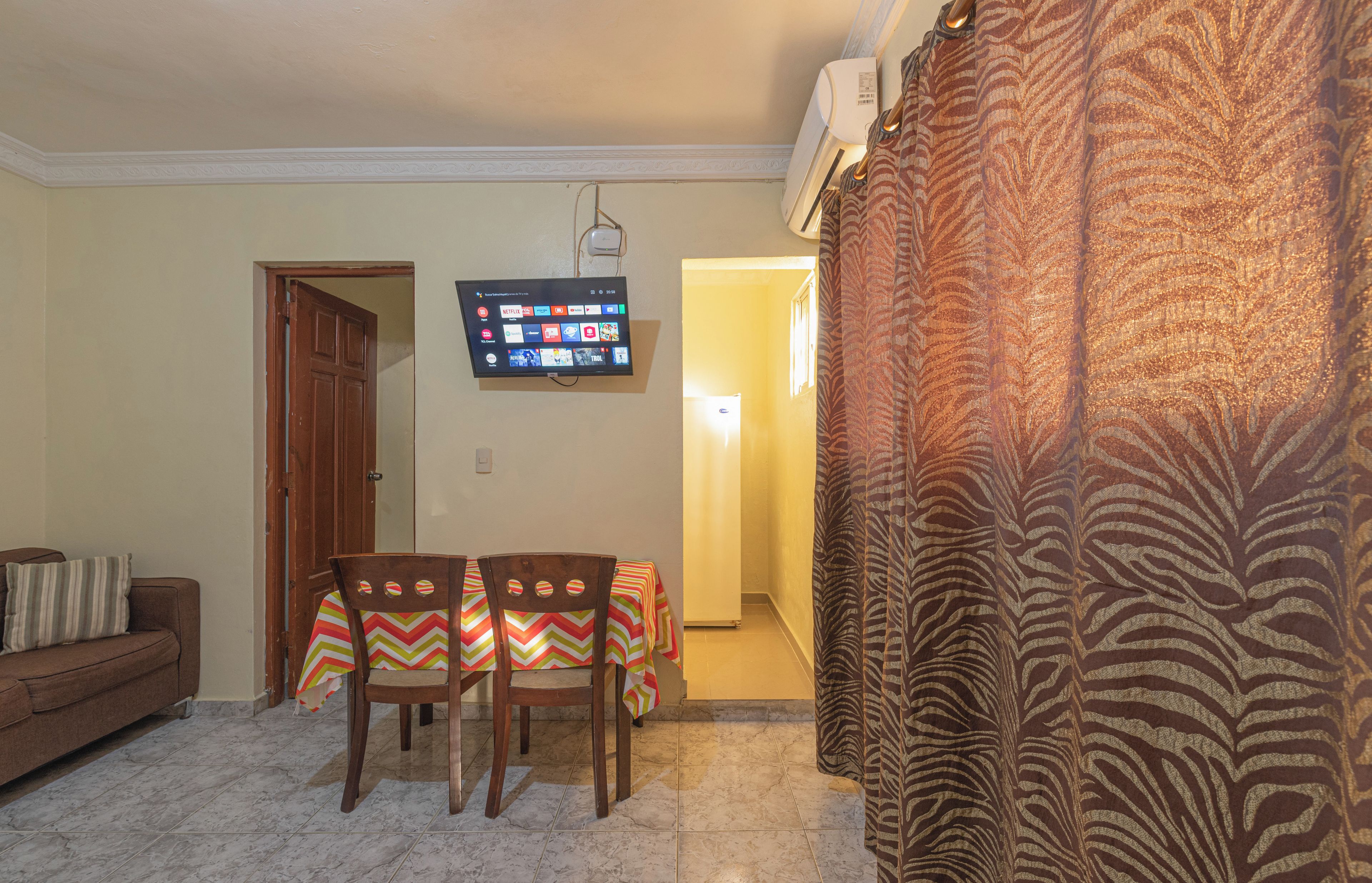 A living room with a sofa, a dining table with chairs, a flat-screen TV, and a patterned curtain.