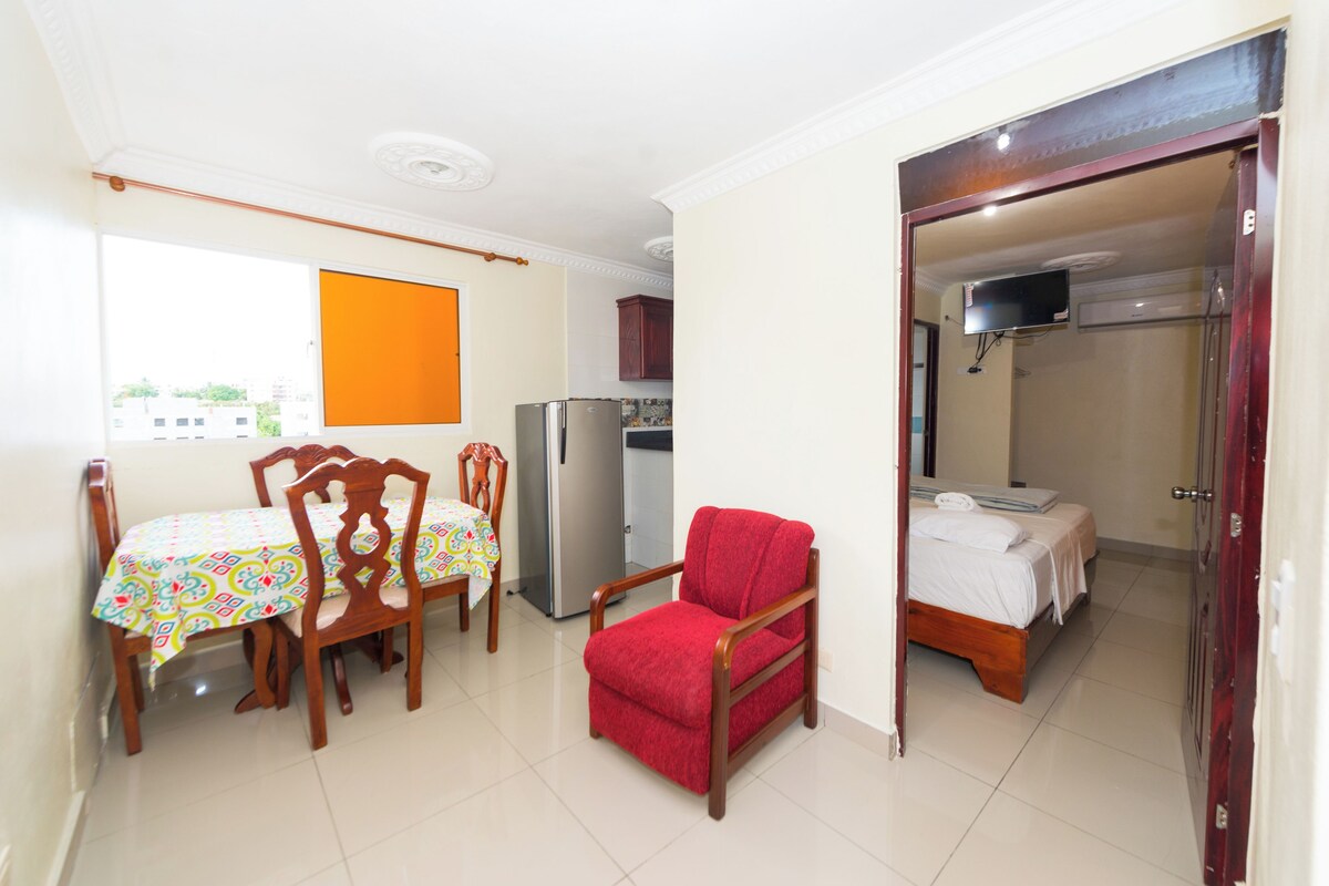A room with a dining table, chairs, a red armchair, a refrigerator, a bed, and a TV.