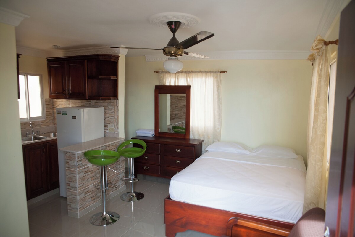 A compact hotel room with a kitchenette, a bed, a bathroom, and a ceiling fan.