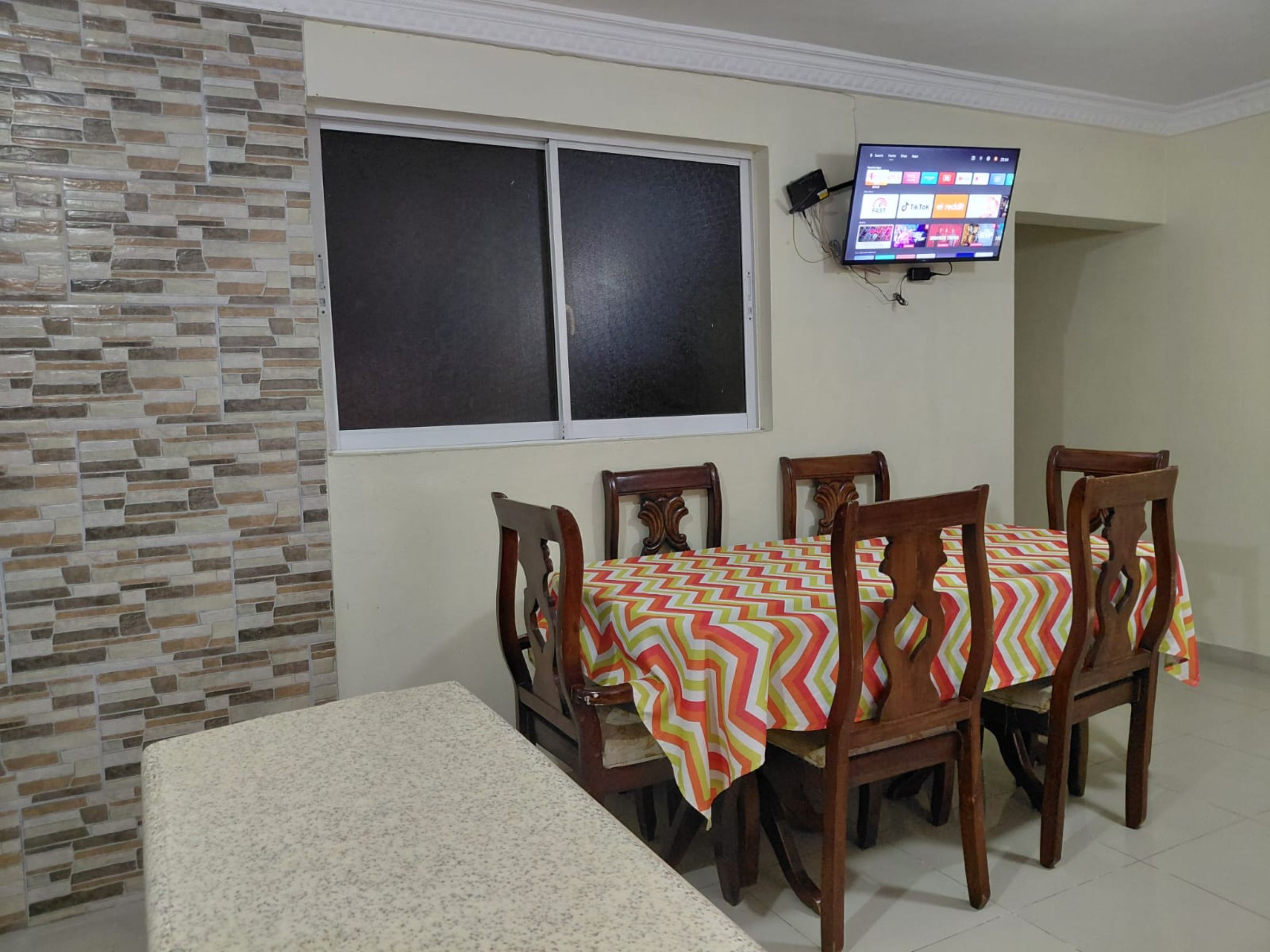 A dining area with a table set for four, a television mounted on the wall, and a stone-patterned wall.