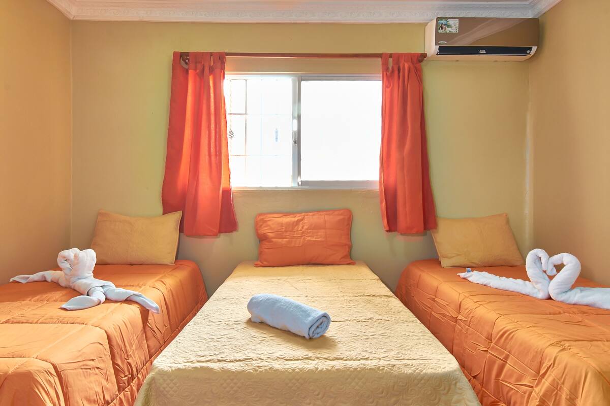 Hotellihuone, jossa on kaksi sänkyä, oranssit vuodevaatteet, keskellä oleva sänky, jonka päällä on rullalle taitettu pyyhe, ja ikkuna, jossa on oranssit verhot.