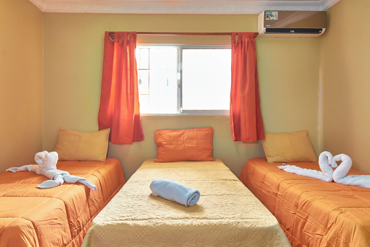A hotel room with two beds, orange bedding, a central bed with a rolled towel, and a window with orange curtains.