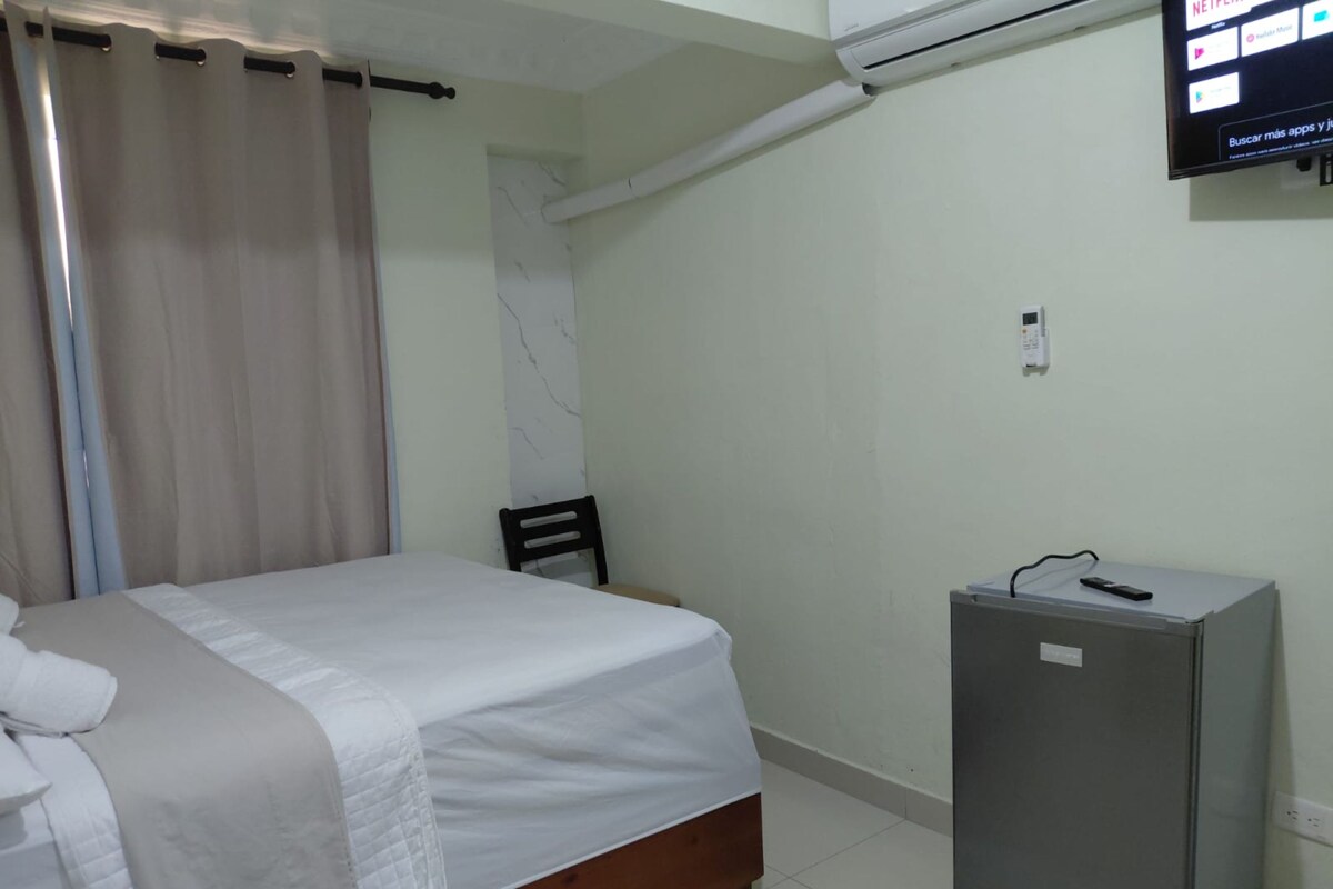 A hotel room with a bed, a small desk, a TV mounted on the wall, and a window with curtains.