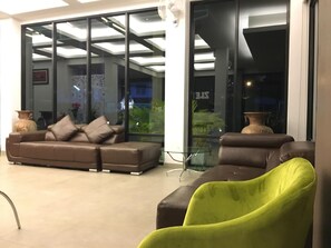 Un espacio de sala moderno con un sofá y sillones, una mesa de vidrio y vistas amplias a través de ventanas grandes.