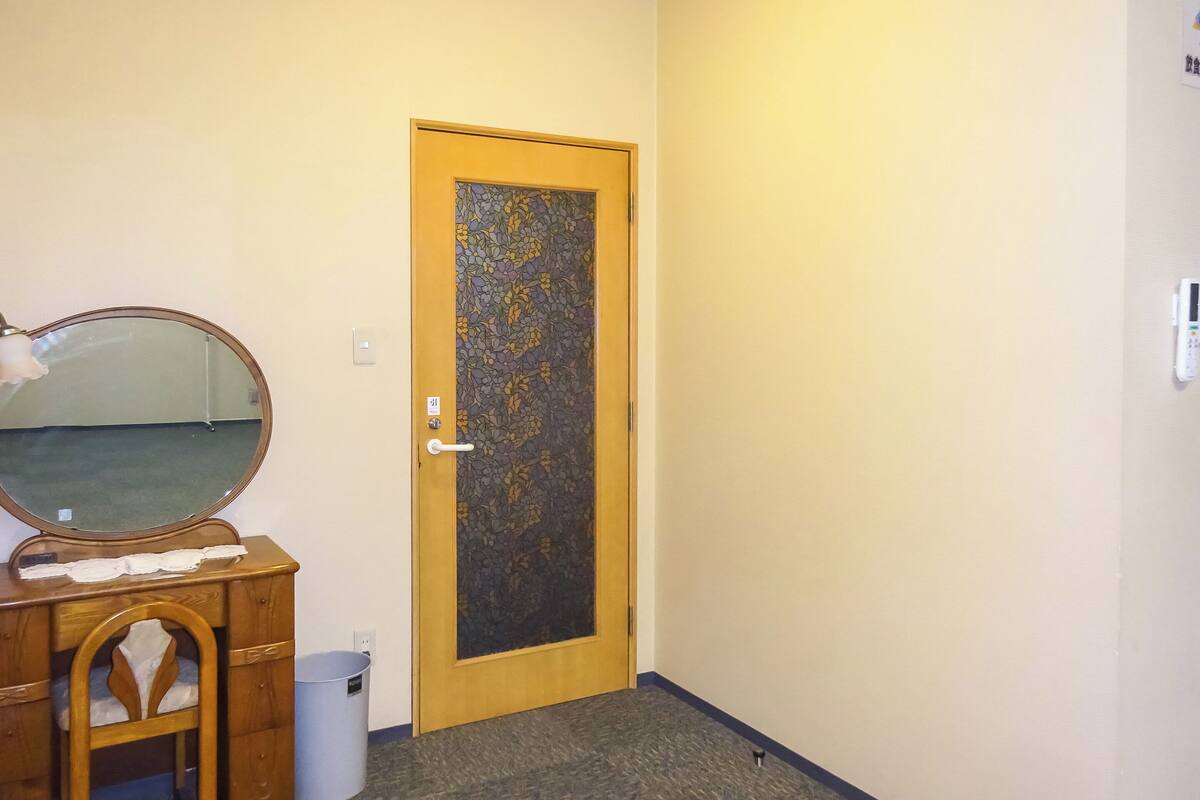 A room with a wooden door, a mirror, a wooden dresser, and a trash bin.