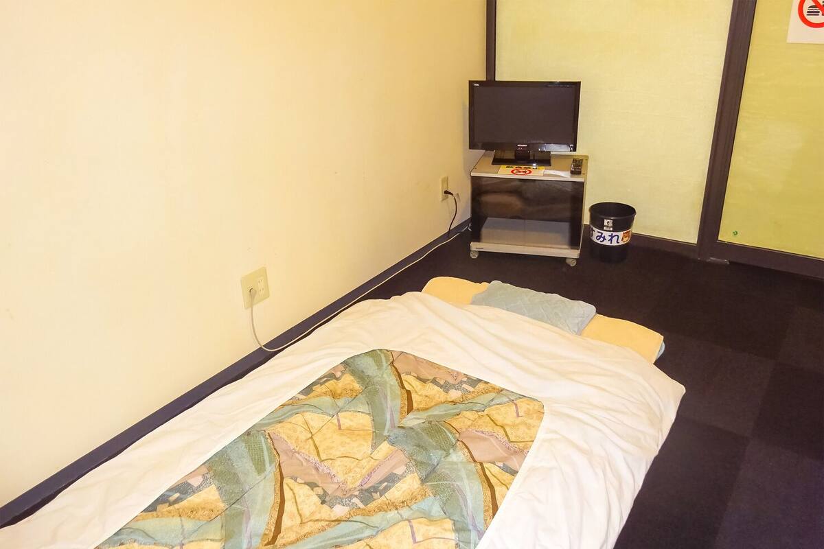 A hotel room with a bed, a television on a stand, a trash bin, and a power outlet on the wall.