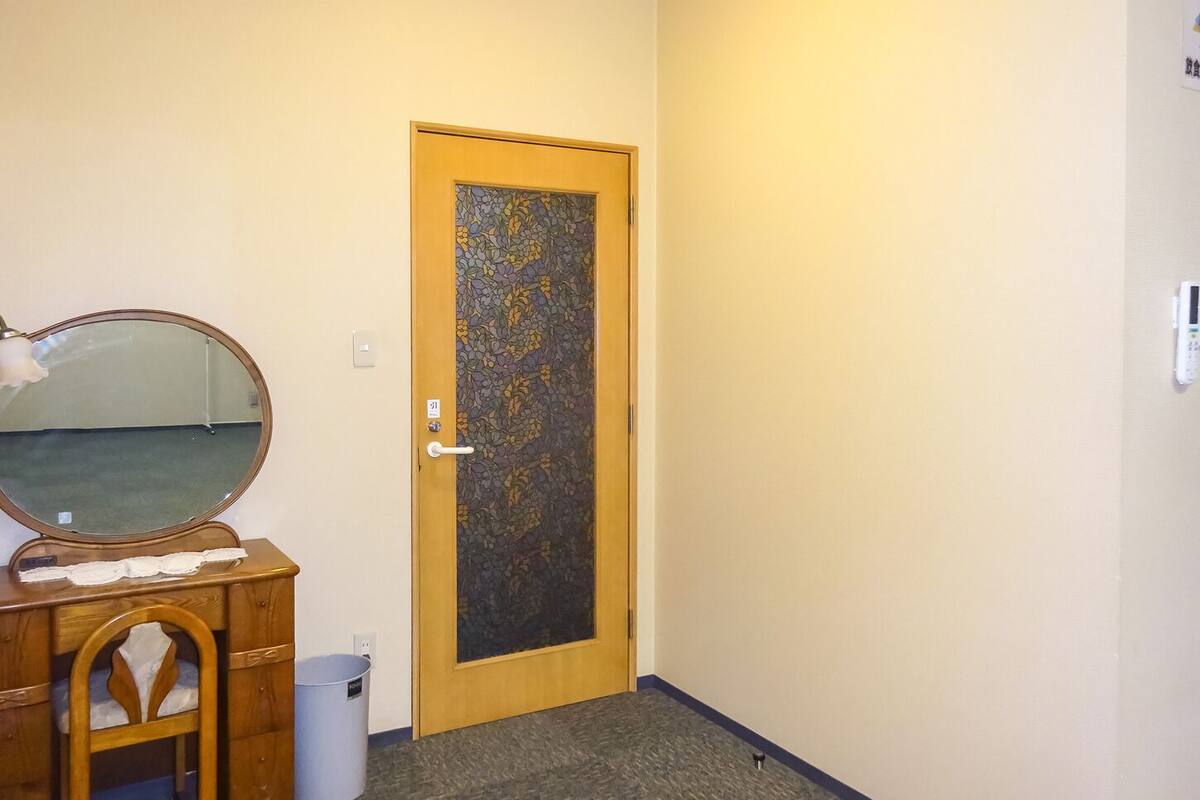A room with a wooden door, a mirror, a wooden dresser, and a trash bin.