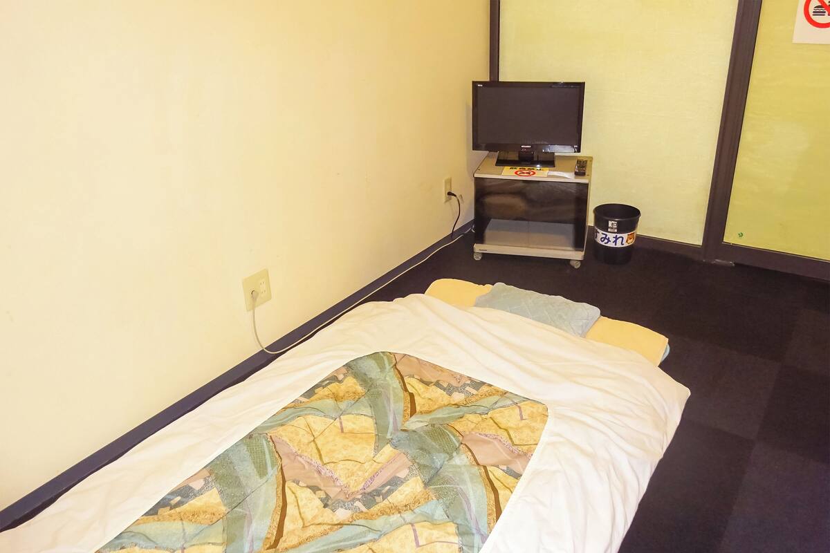 A single bed with a patterned bedspread, a television on a stand, a trash bin, and a power outlet on the wall.