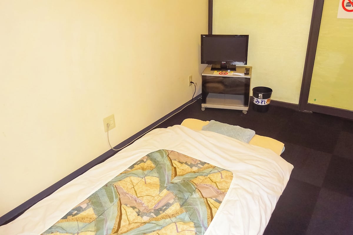 Una cama individual con una colcha estampada, un televisor sobre un mueble, un cubo de basura y un enchufe en la pared.