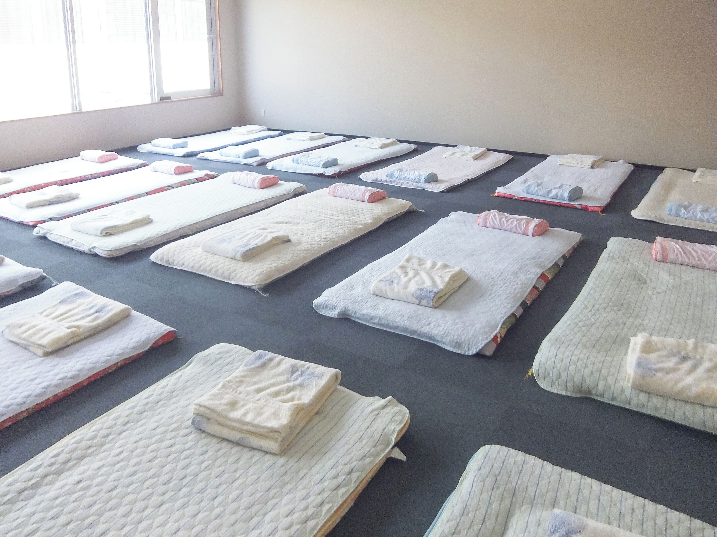 Una habitación con varias camas dispuestas en filas, cada una con una manta y almohada dobladas con cuidado.