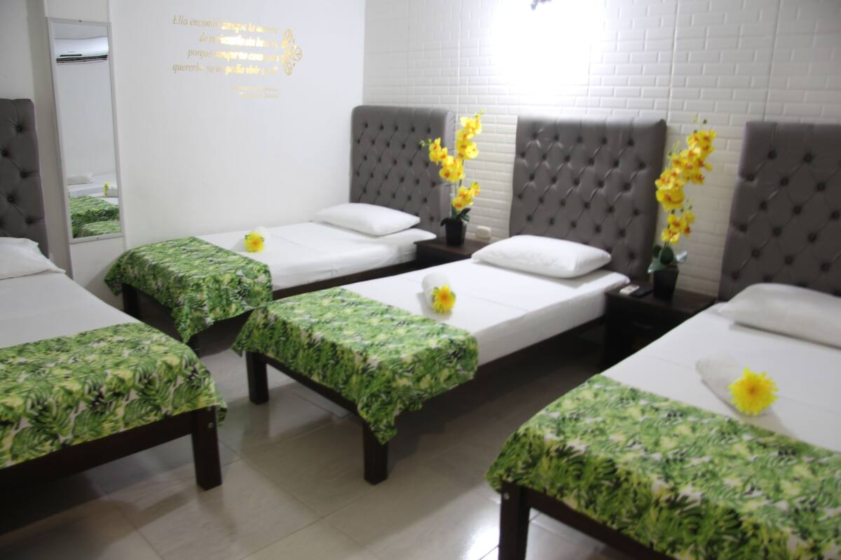 A hotel room with three single beds, each with a green floral bedspread, a mirror, and a wall with a white brick pattern.