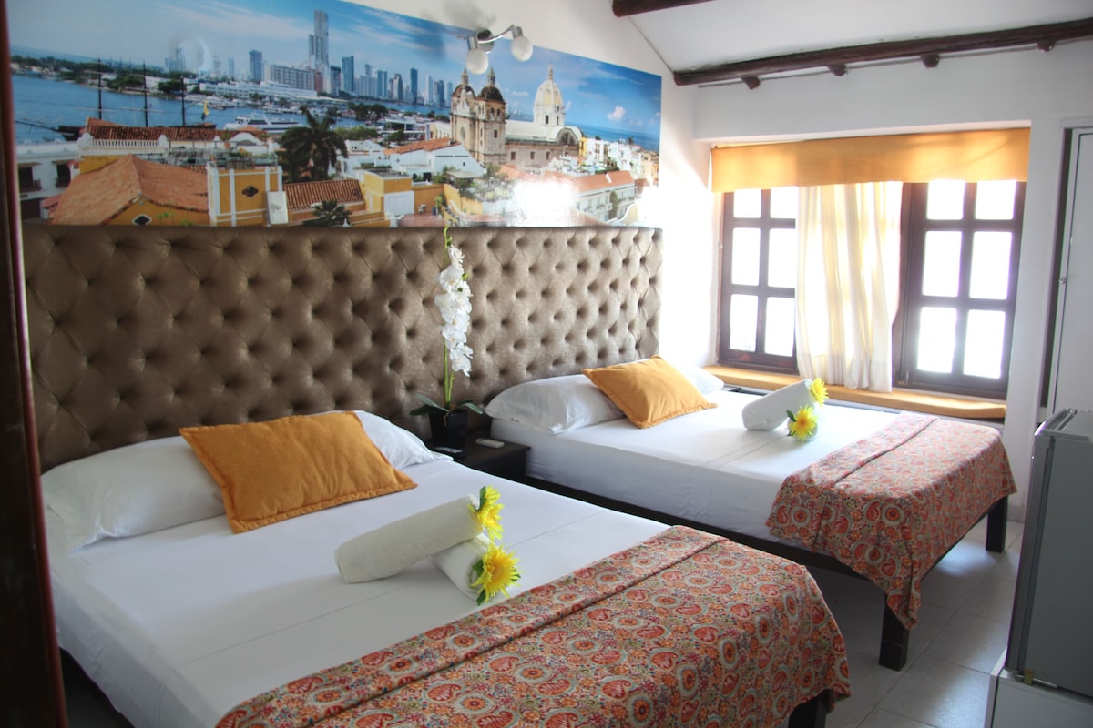 A hotel room with two beds, a large cityscape mural, a window with curtains, and a view of the city.