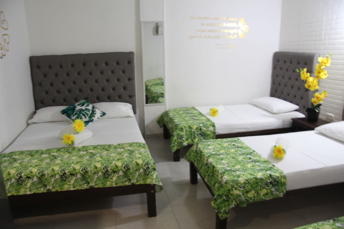 A hotel room with three beds, each with a green leaf-patterned bedspread, a grey tufted headboard, and a white pillow.