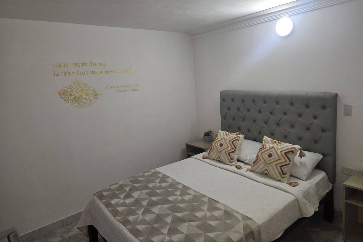 A bedroom with a grey tufted headboard, a patterned bedspread, and a wall with a decorative quote and feather design.