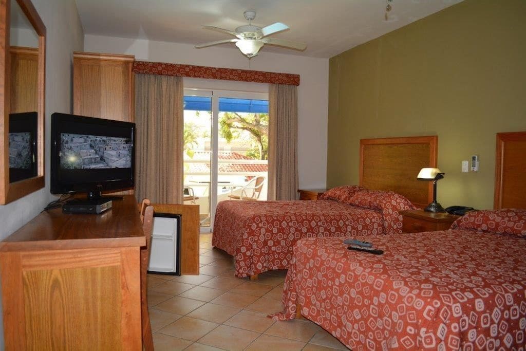 A hotel room with a television, two beds with patterned bedding, a ceiling fan, and a view of the outdoors.