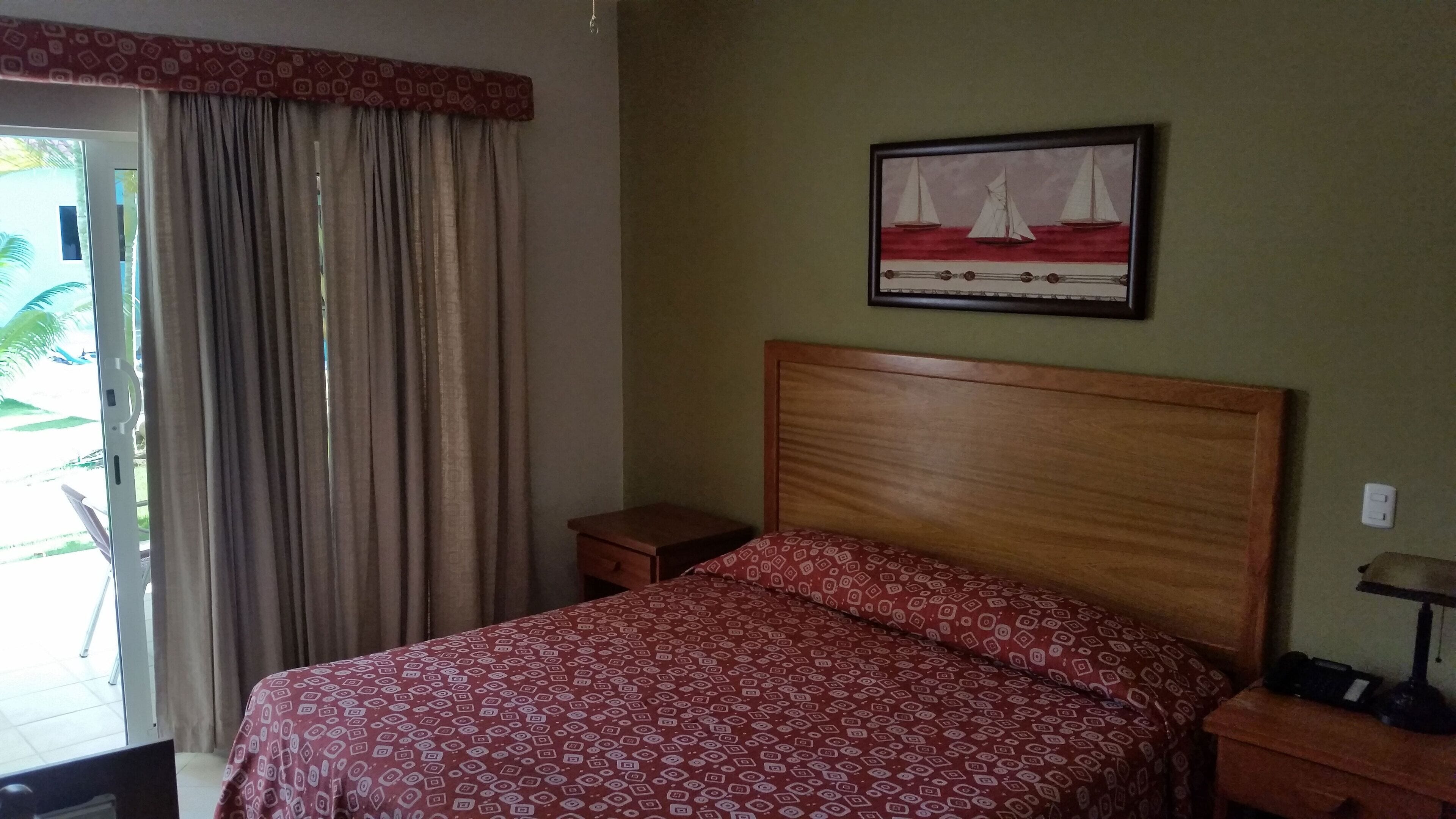 Un dormitorio con cama, mesita de noche, lámpara, cuadro y ventana con cortinas.