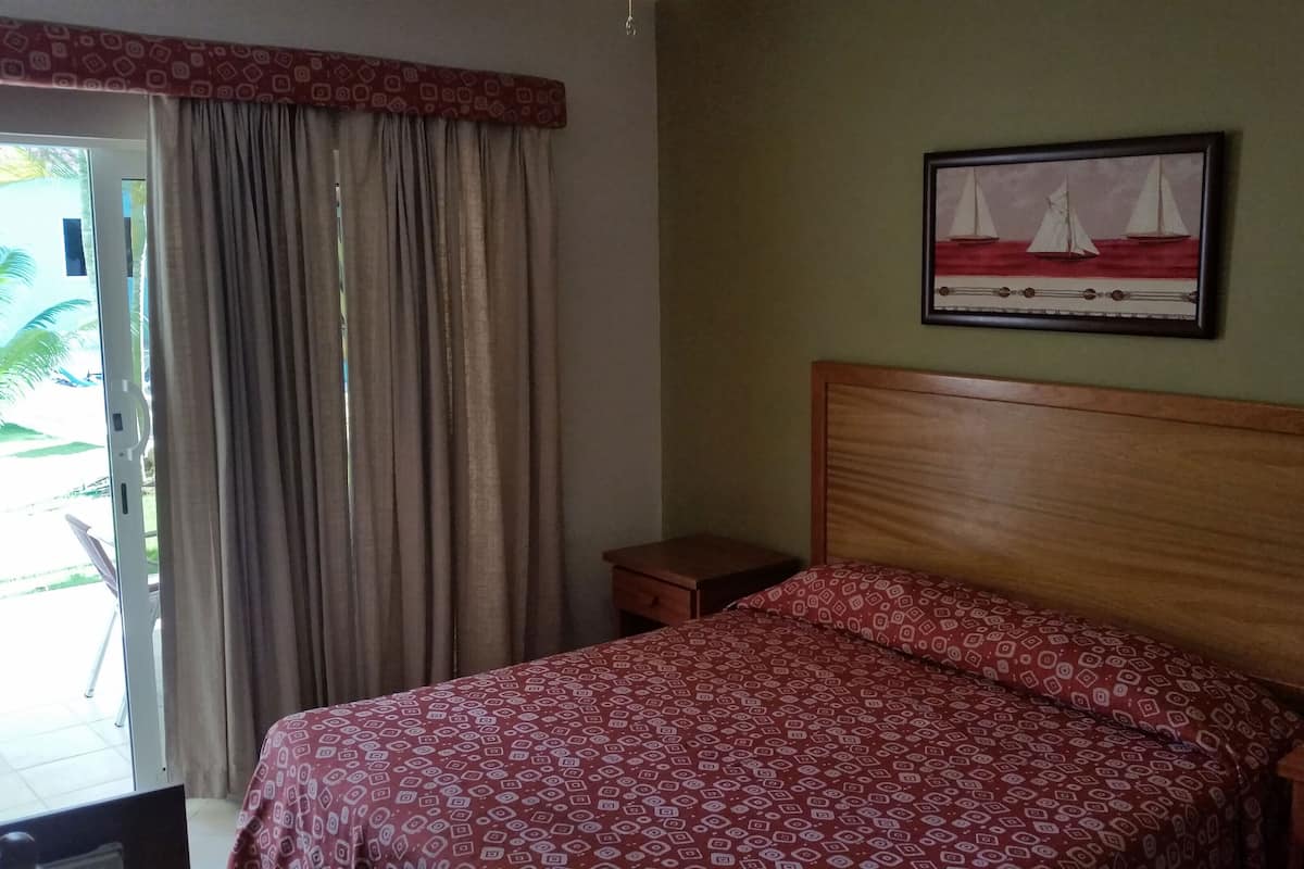 Un dormitorio con cama, mesita de noche, lámpara, cuadro y ventana con cortinas.