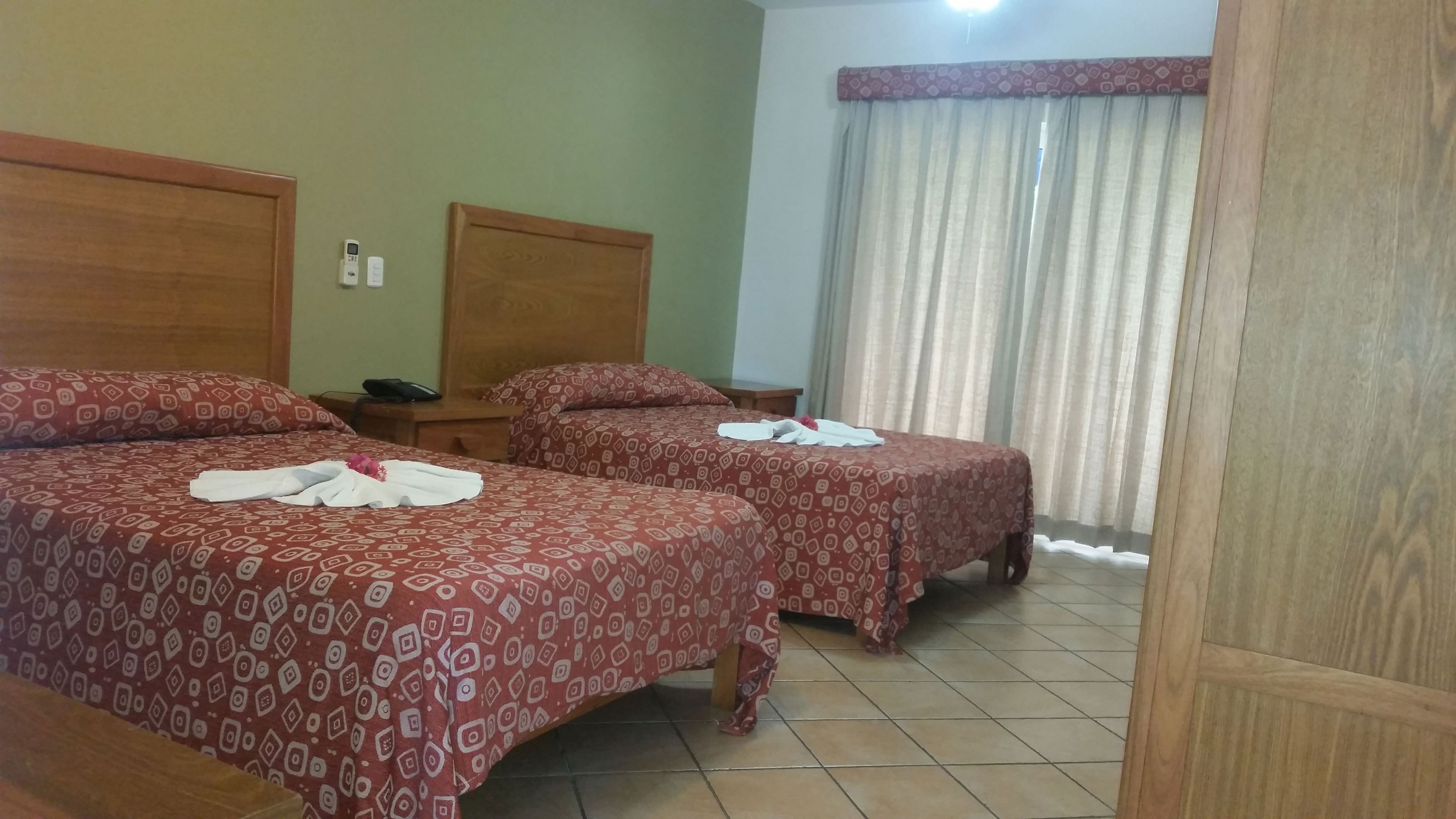 A hotel room with two beds, each with patterned bedding and towels. There is a nightstand with a phone and a wall-mounted thermostat.