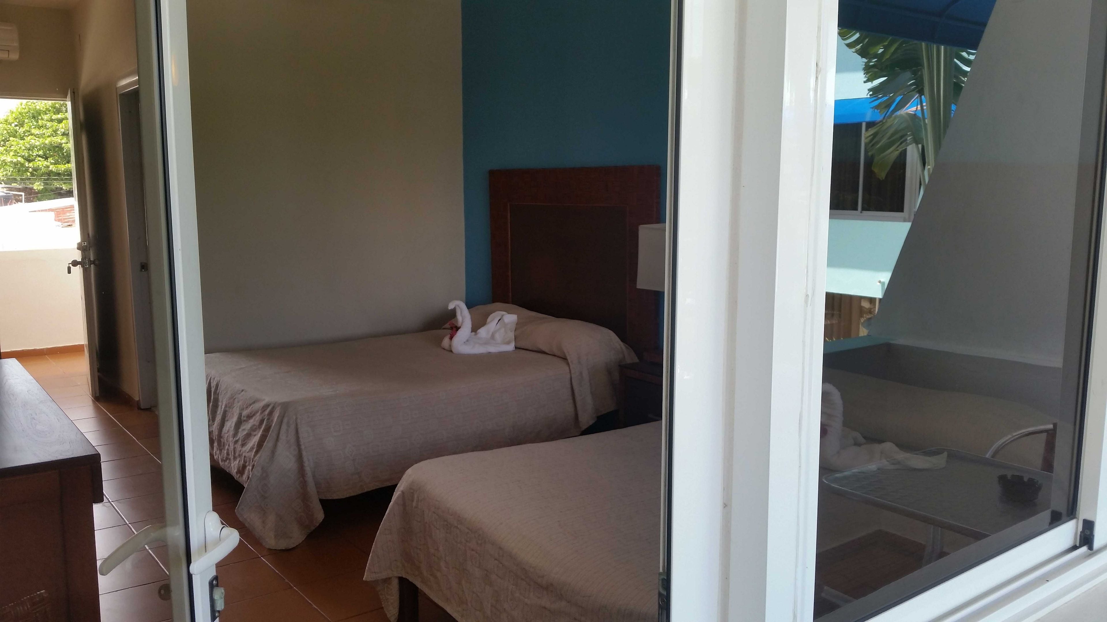 Quarto de hotel com duas camas, cabeceira de madeira, janela com vista para palmeiras e porta que dá para uma varanda.