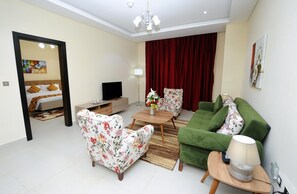 Ένα δωμάτιο ξενοδοχείου με ένα κρεβάτι, μια τηλεόραση, μια πολυθρόνα με σχέδια λουλουδιών, έναν πράσινο καναπέ και ένα μικρό τραπέζι με ένα φωτιστικό.