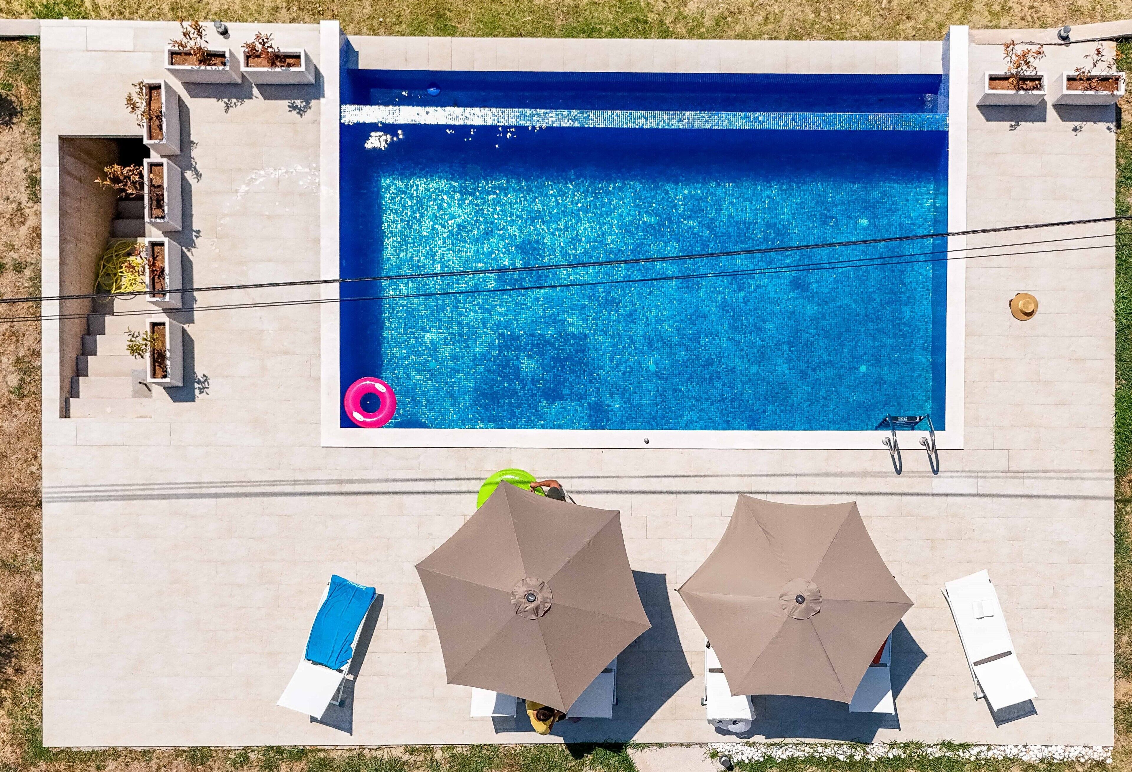 A rectangular swimming pool with a clear blue water surface, surrounded by a light-colored tiled deck. There are two umbrellas and lounge chairs, and a small section of the deck has planters with greenery.
