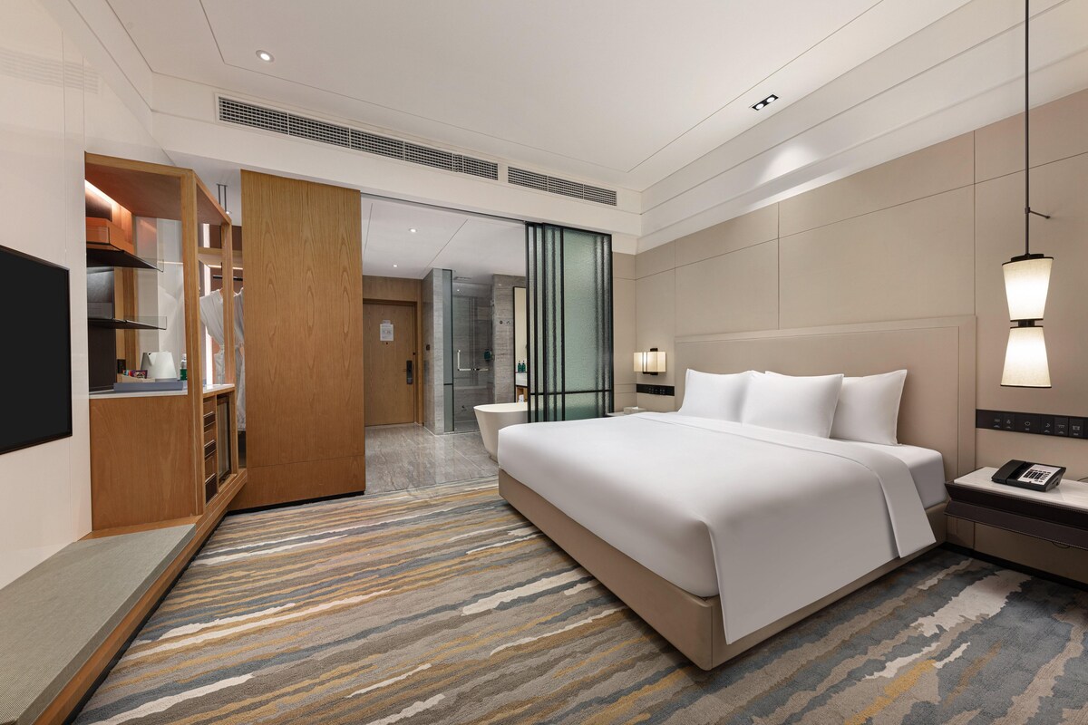 A modern hotel room with a large bed, a walk-in closet, a bathroom with a glass shower, and a seating area with a bench.