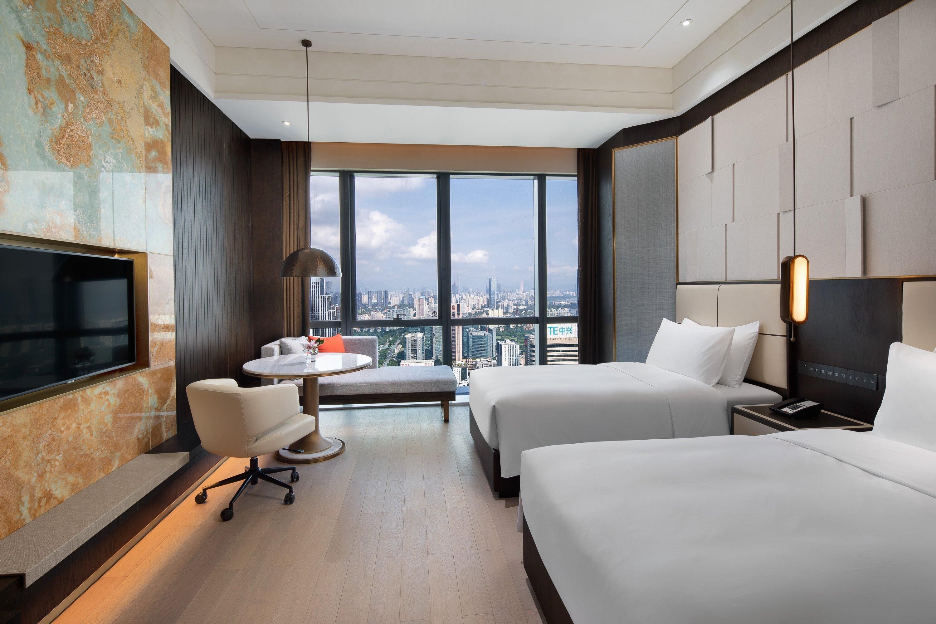 Ein Hotelzimmer mit zwei Betten, einem Fernseher, einem Schreibtisch und einem großen Fenster mit Blick auf die Stadt.