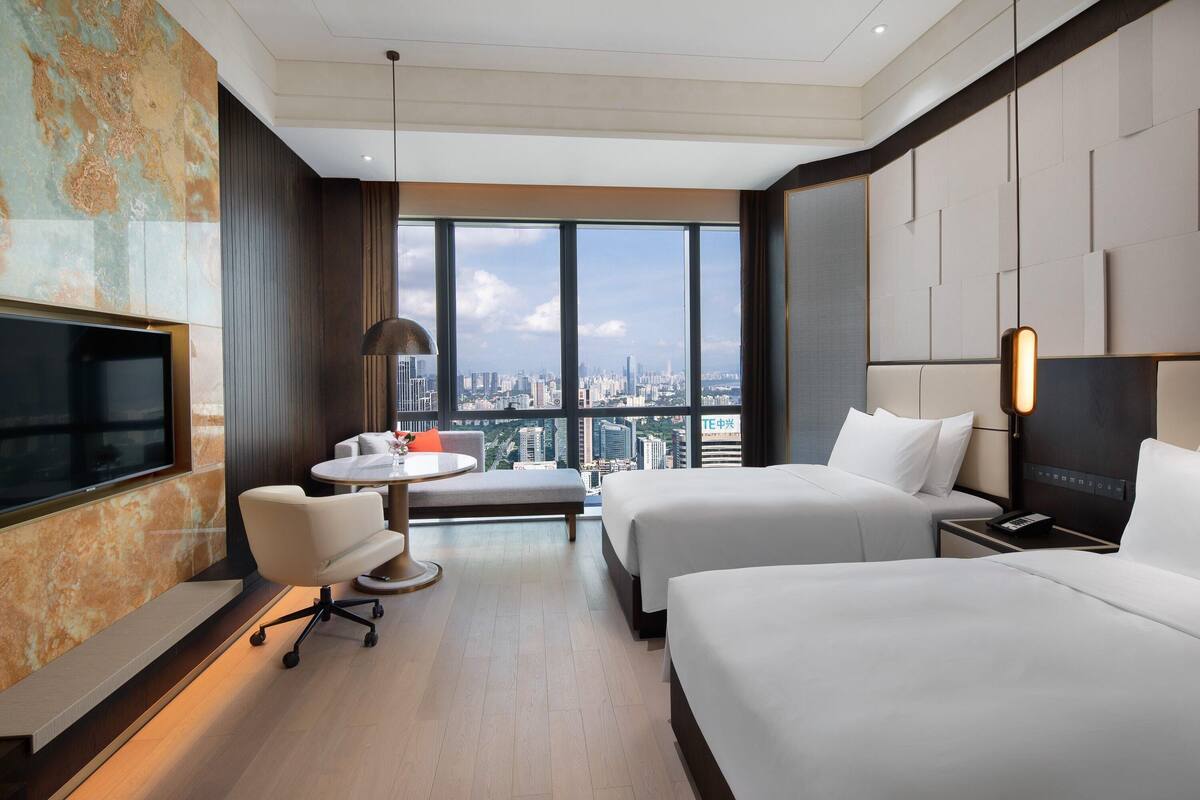 Quarto de hotel com duas camas, televisão, escrivaninha e uma grande janela com vista para a cidade.
