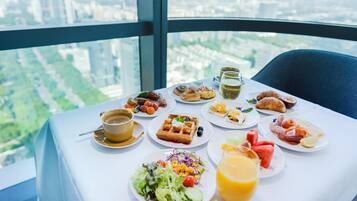 Mesa com itens de café da manhã, incluindo waffles, croissants e diversas frutas, com vista para a cidade.