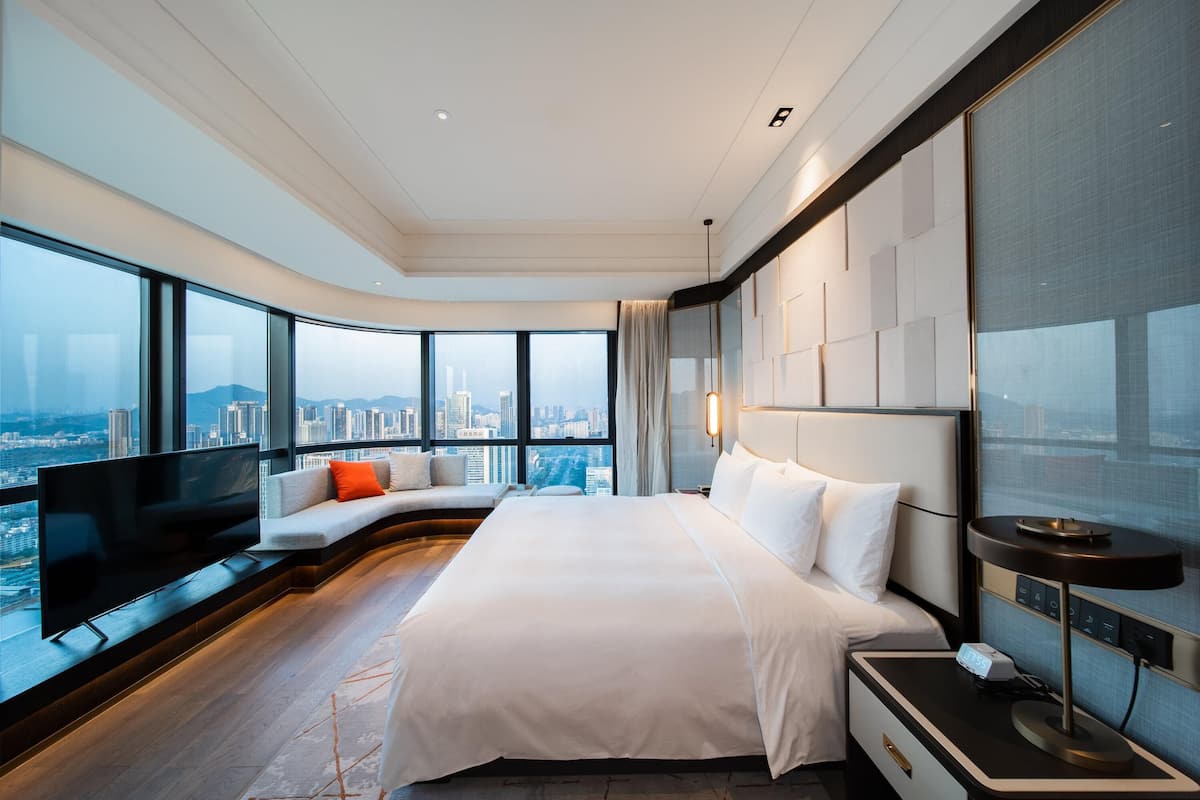 Quarto de hotel moderno com uma cama grande, televisão, sofá e vista para a cidade.