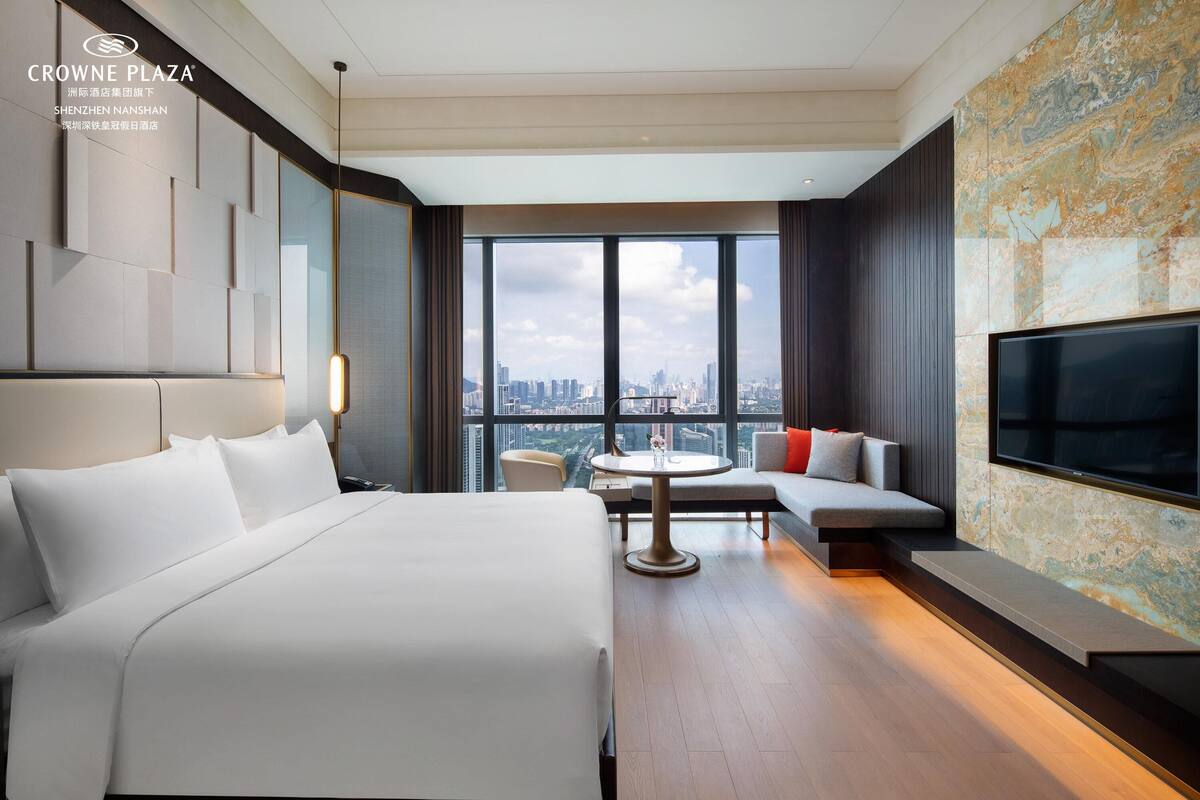Quarto de hotel moderno com uma cama grande, área de estar com uma mesa e vista para a cidade através de janelas amplas.