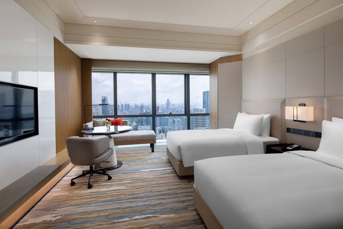 Quarto de hotel com duas camas, uma TV, uma escrivaninha com cadeira, uma mesinha com um vaso vermelho e vista para a cidade através de grandes janelas.