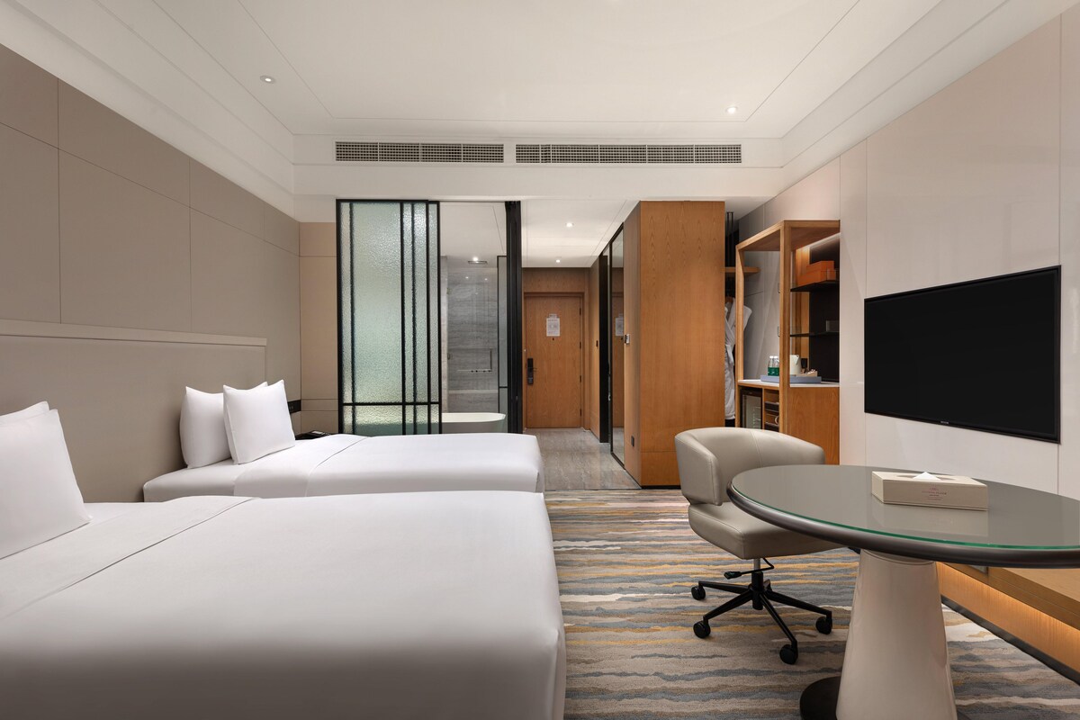 A hotel room with two beds, a desk, a chair, a TV, and a bookshelf.