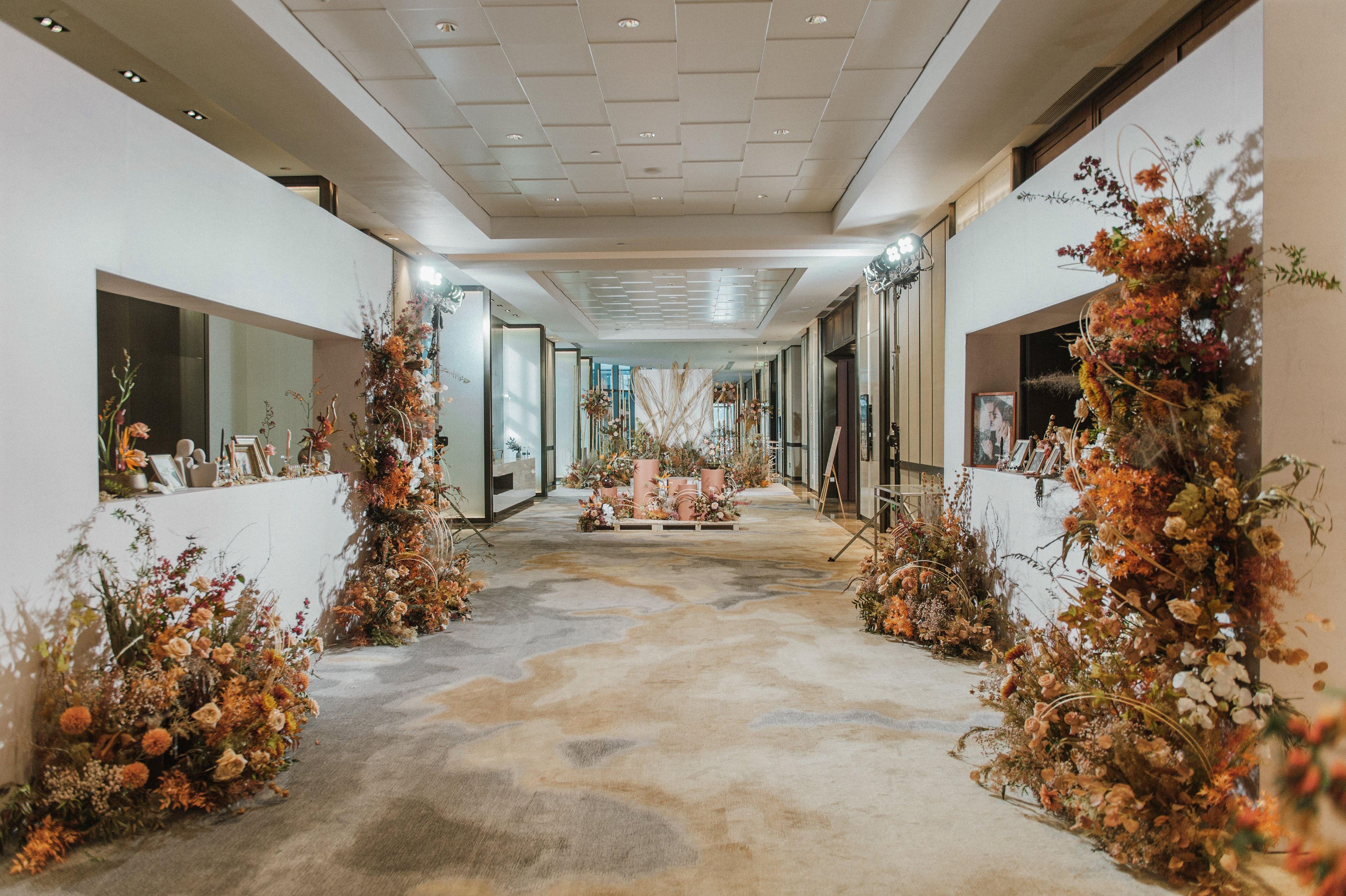 Un pasillo con un arreglo floral central, flanqueado por paredes con cuadros enmarcados y plantas decorativas.