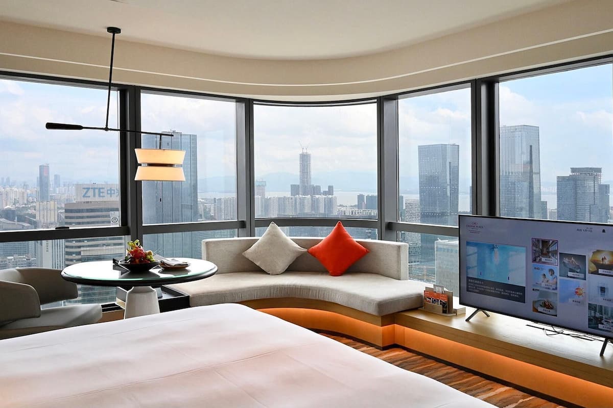 Quarto de hotel moderno com teto curvo, cama grande, sofá com almofadas, mesa pequena com uma cesta de frutas e TV exibindo uma vista da cidade.