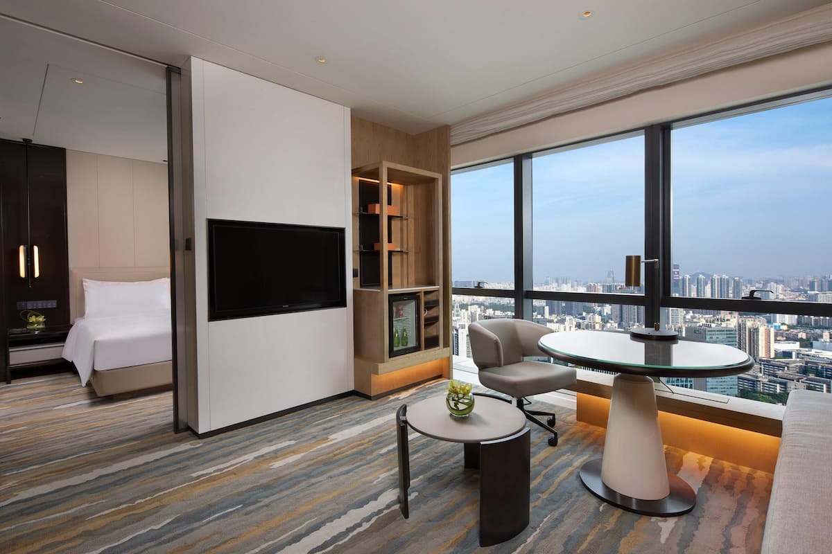 Quarto de hotel moderno com cama, TV, área de estar com mesa e uma janela grande com vista para a cidade.