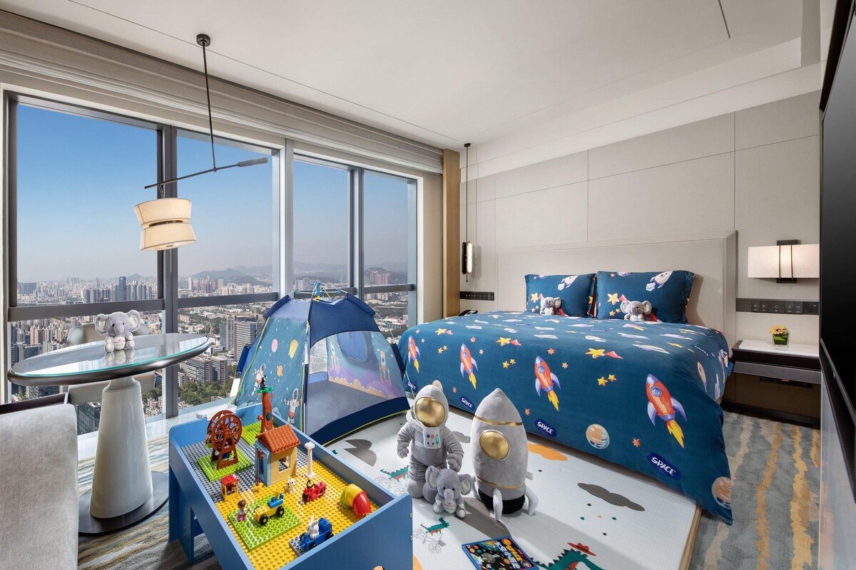 A modern hotel room with a large window offering a city view, a bed with space-themed bedding, a play area with toys, and a small table.