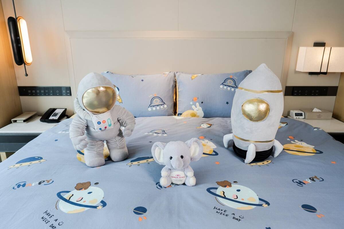 Quarto de hotel com uma cama, um edredom com tema espacial, dois travesseiros com tema de astronauta e um pequeno brinquedo de pelúcia.