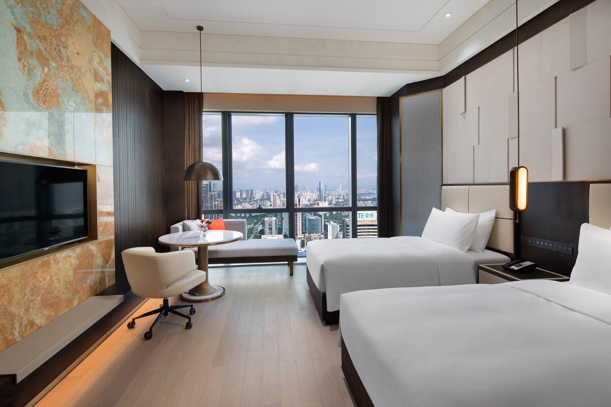 A hotel room with two beds, a TV, a desk, and a large window with a city view.