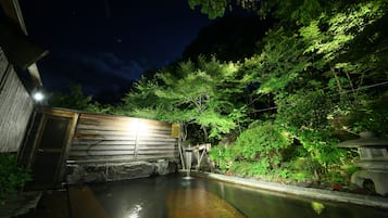 Vista nocturna de un onsen japonés tradicional con pared de madera, sendero de piedra y exuberante vegetación.