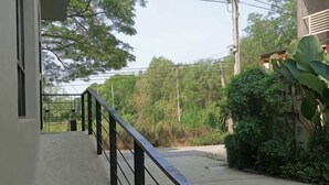 Uma varanda com guarda-corpo de metal, piso de concreto e vista para o verde e uma estrada.