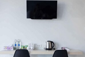 Una habitación con una televisión de pantalla plana montada en la pared, un escritorio de color claro con un hervidor, un teléfono y algunas botellas.