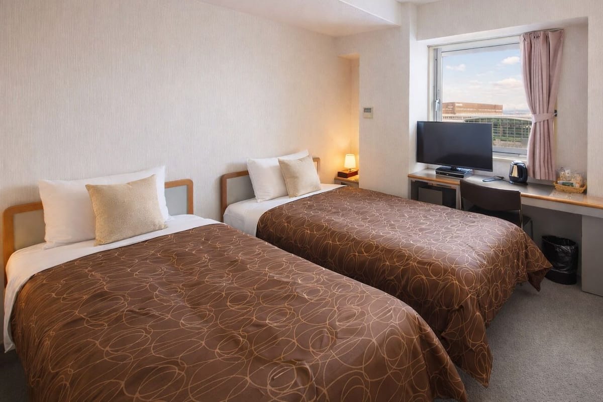 A hotel room with two beds, a desk with a TV, a window with a view, and a lamp.