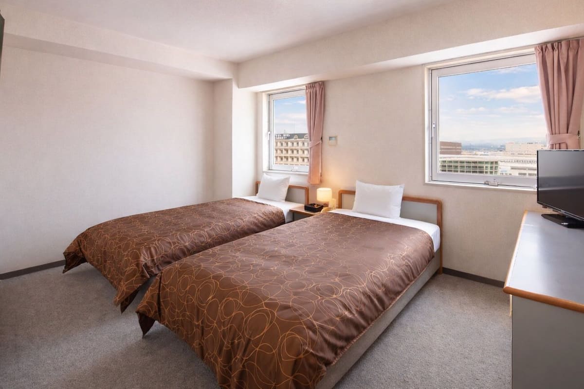 A hotel room with two beds, a TV, a desk, and a window with a city view.